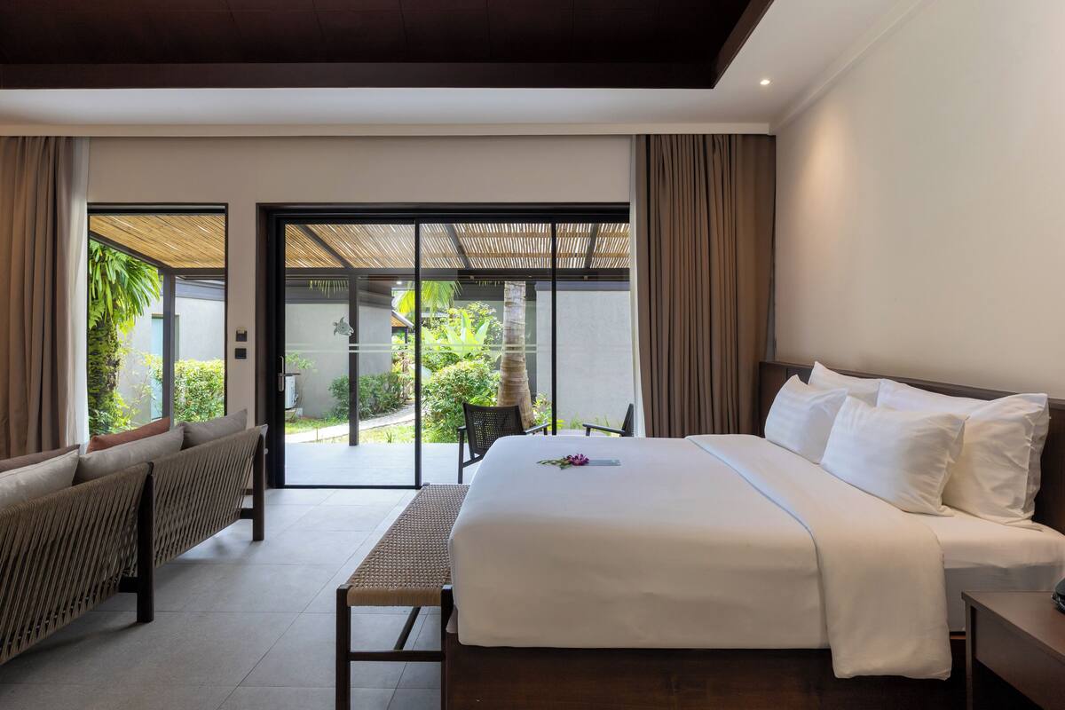 Una habitación de hotel moderna con una cama grande, una zona de estar con un banco y vistas a un jardín a través de una puerta corredera.