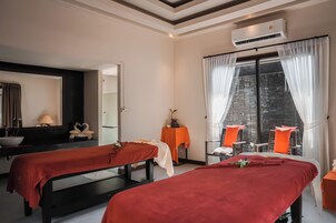 Un cuarto de spa con dos camillas de masaje, una pared de piedra, una ventana con cortinas y una pequeña mesa con un florero.