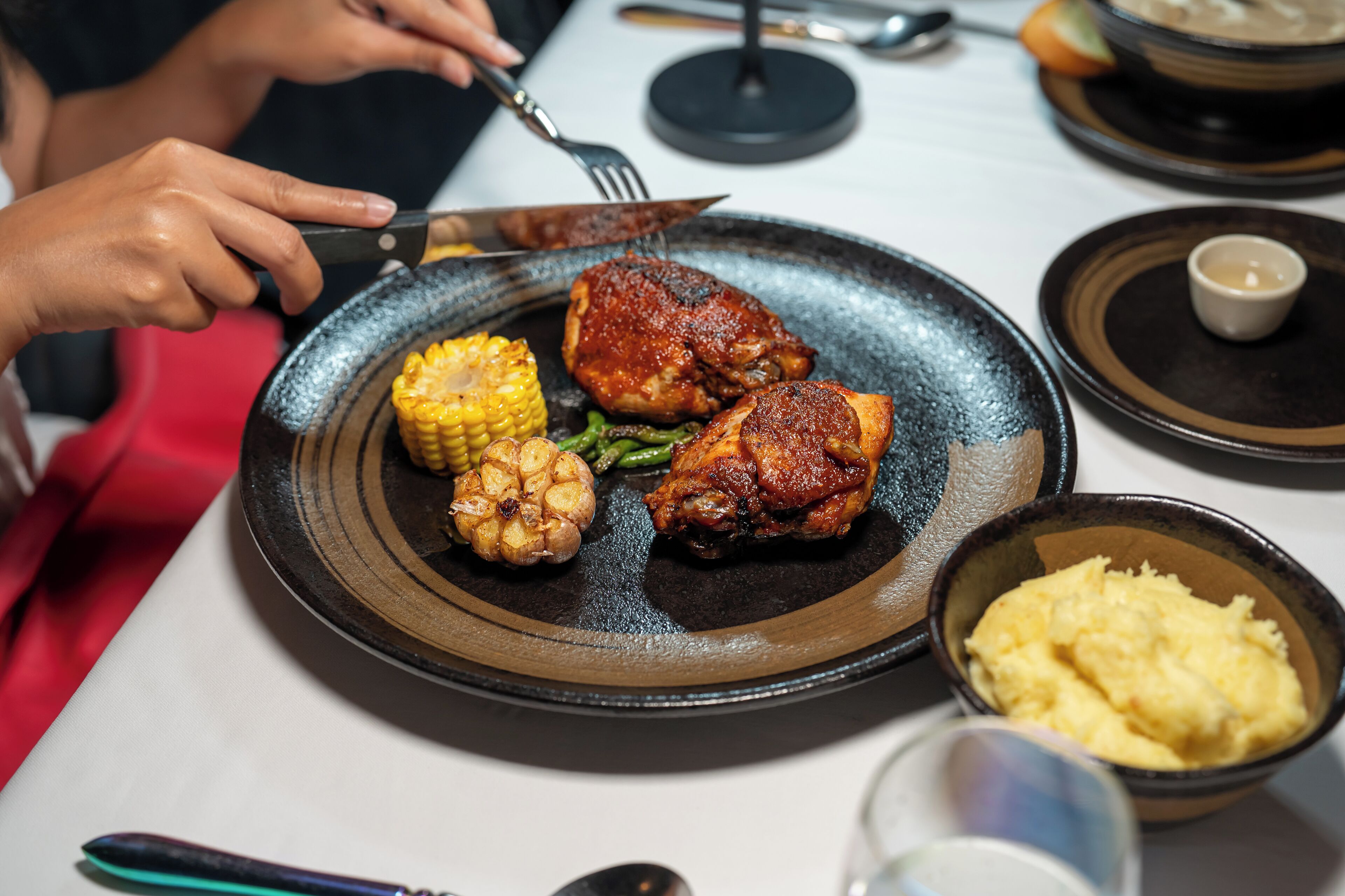 Un plato con pollo a la parrilla, maíz y verduras, un bol de puré de papas y un vaso de agua sobre una mesa.