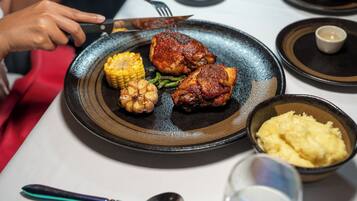 Un plato con pollo a la parrilla, maíz y verduras, un bol de puré de papas y un vaso de agua sobre una mesa.