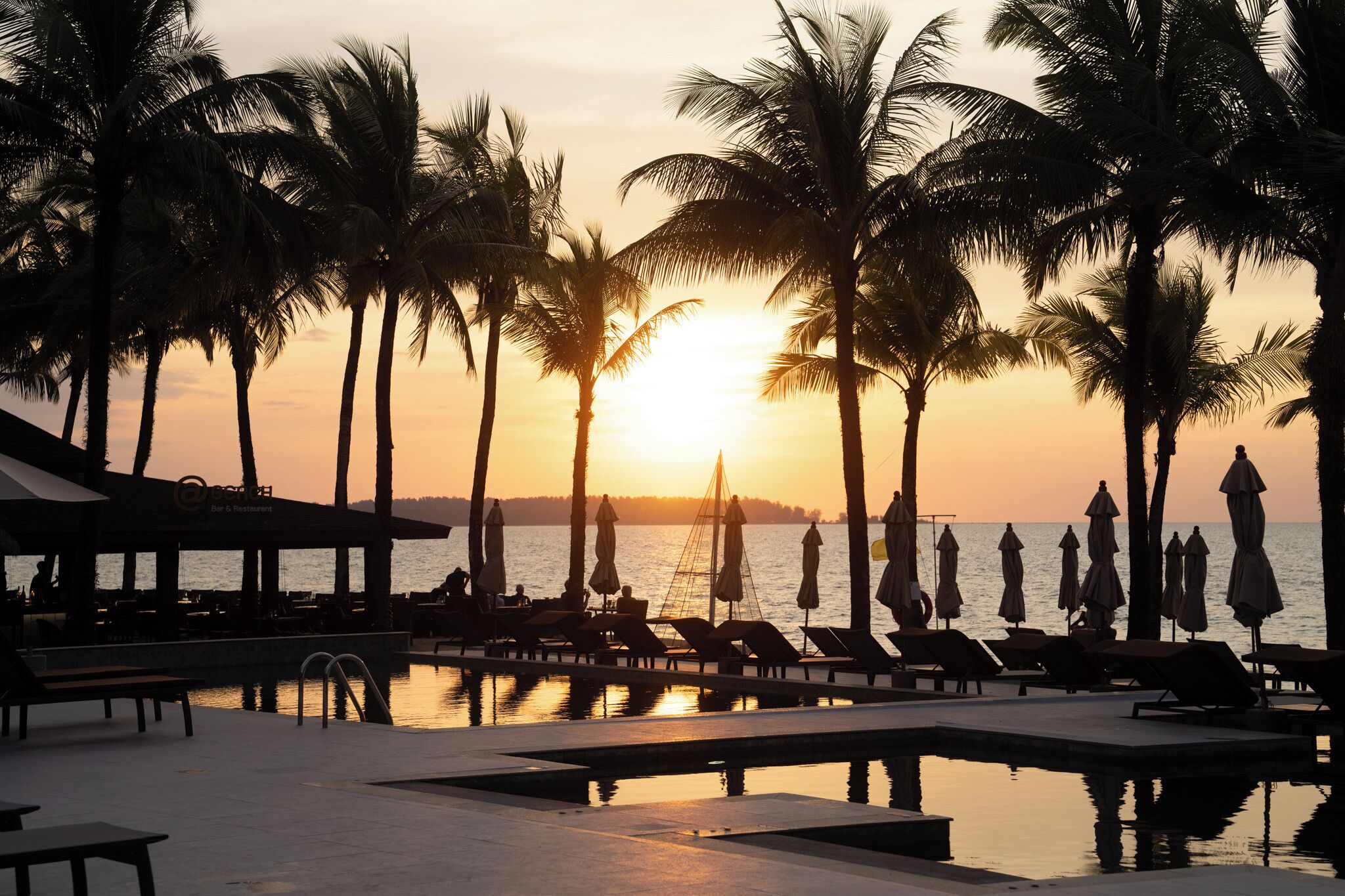 Un área junto a la piscina con sillones de descanso y sombrillas, palmeras y vistas al atardecer.