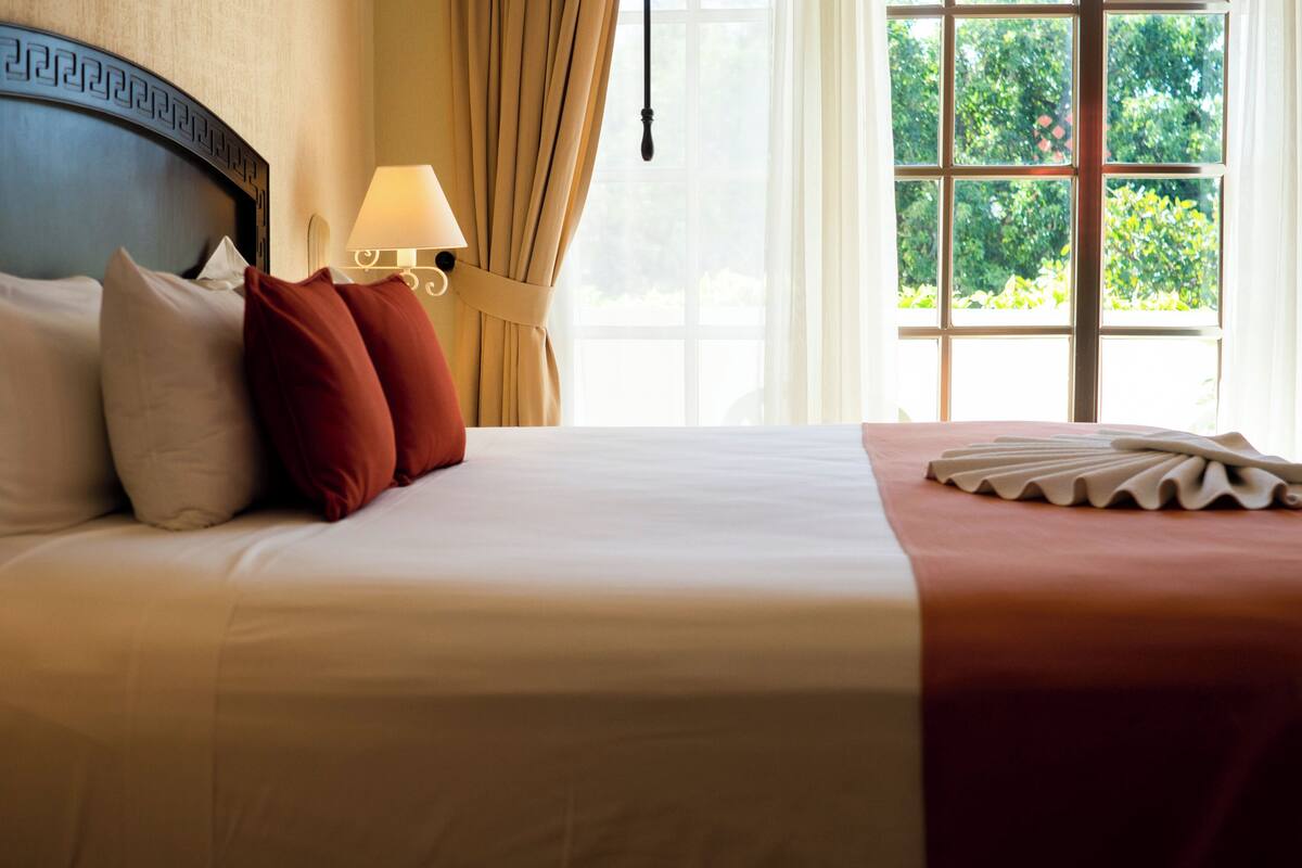A neatly made bed with white and red pillows, a bedside lamp, and a view of greenery through a window.