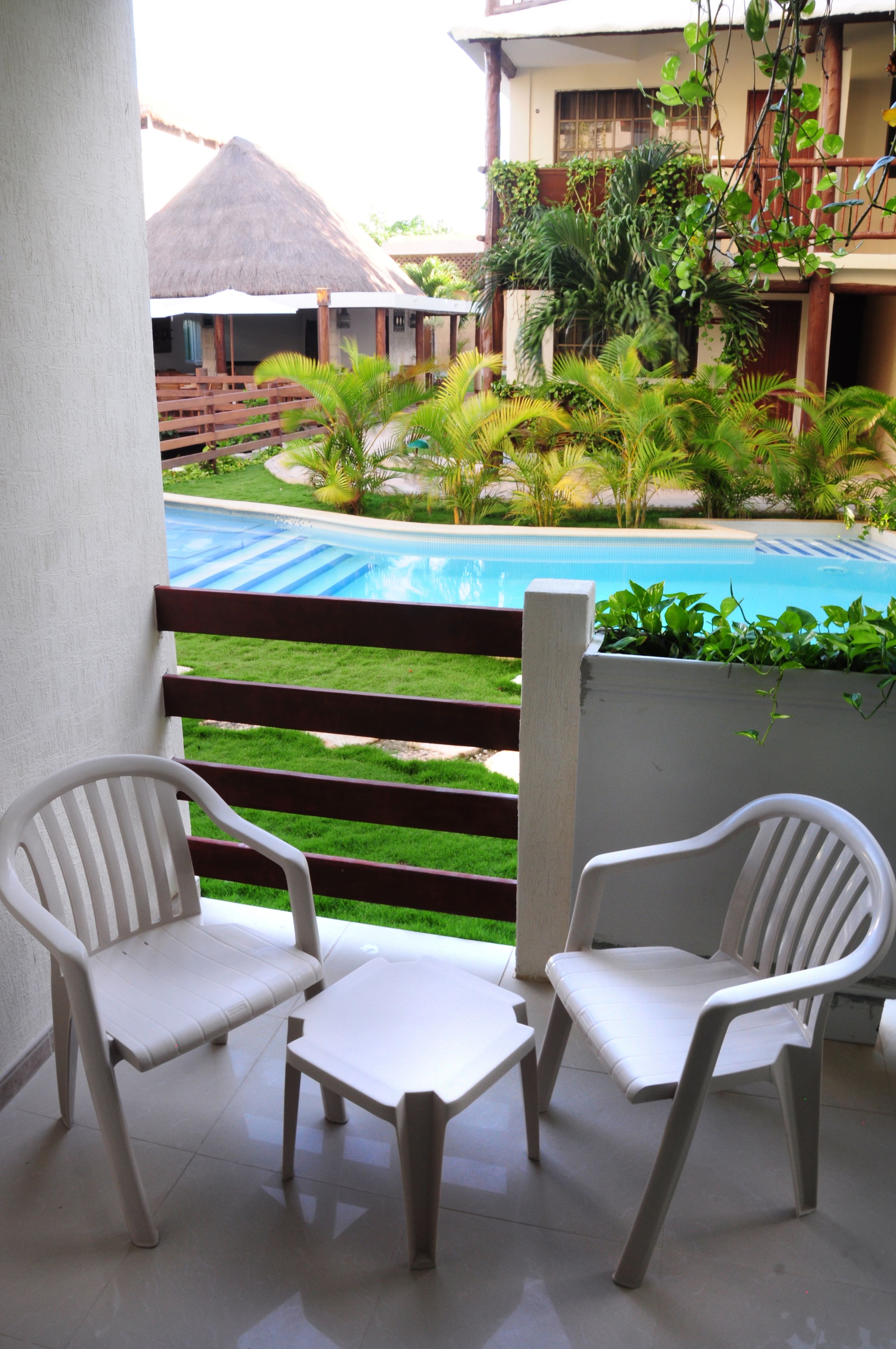 Un balcón con dos sillas de plástico blanco y una mesita pequeña, con vista a una piscina y abundante vegetación.