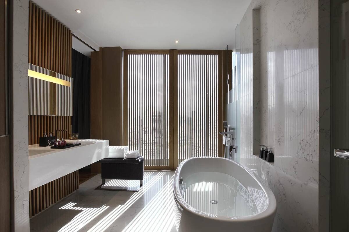 Une salle de bain moderne avec une baignoire sur pieds, un meuble-lavabo avec un miroir et une grande fenêtre dotée de stores verticaux.
