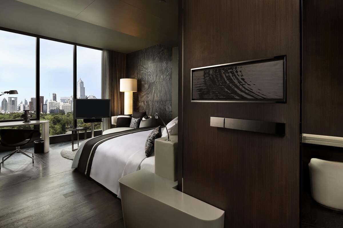 Una habitación de hotel moderna con un ventanal grande, una cama, un escritorio, una silla, una lámpara y vistas a la ciudad.