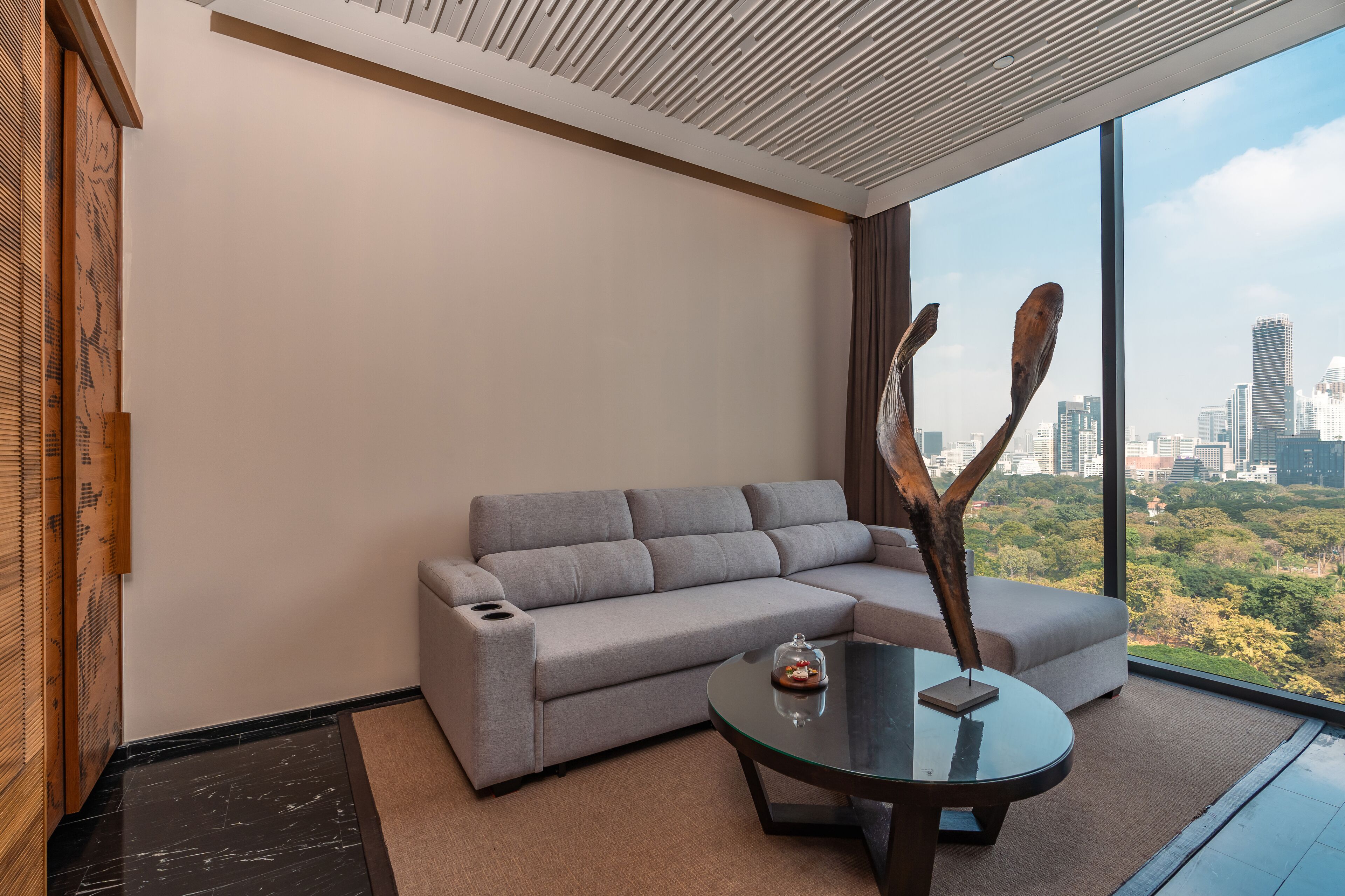 Ein modernes Wohnzimmer mit einem grauen Ecksofa, einem Glastisch und weitreichender Stadtansicht durch große Fenster.