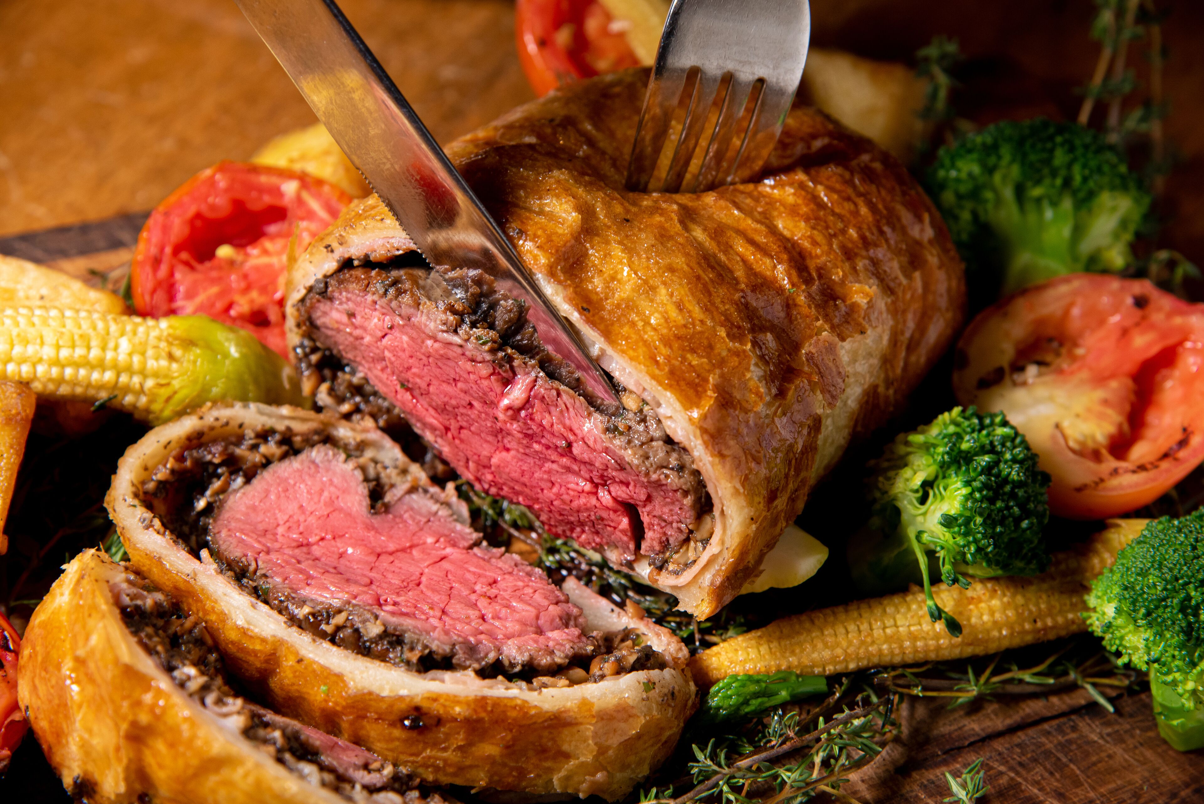 A sliced beef Wellington with a knife cutting through it, surrounded by vegetables like broccoli and corn.