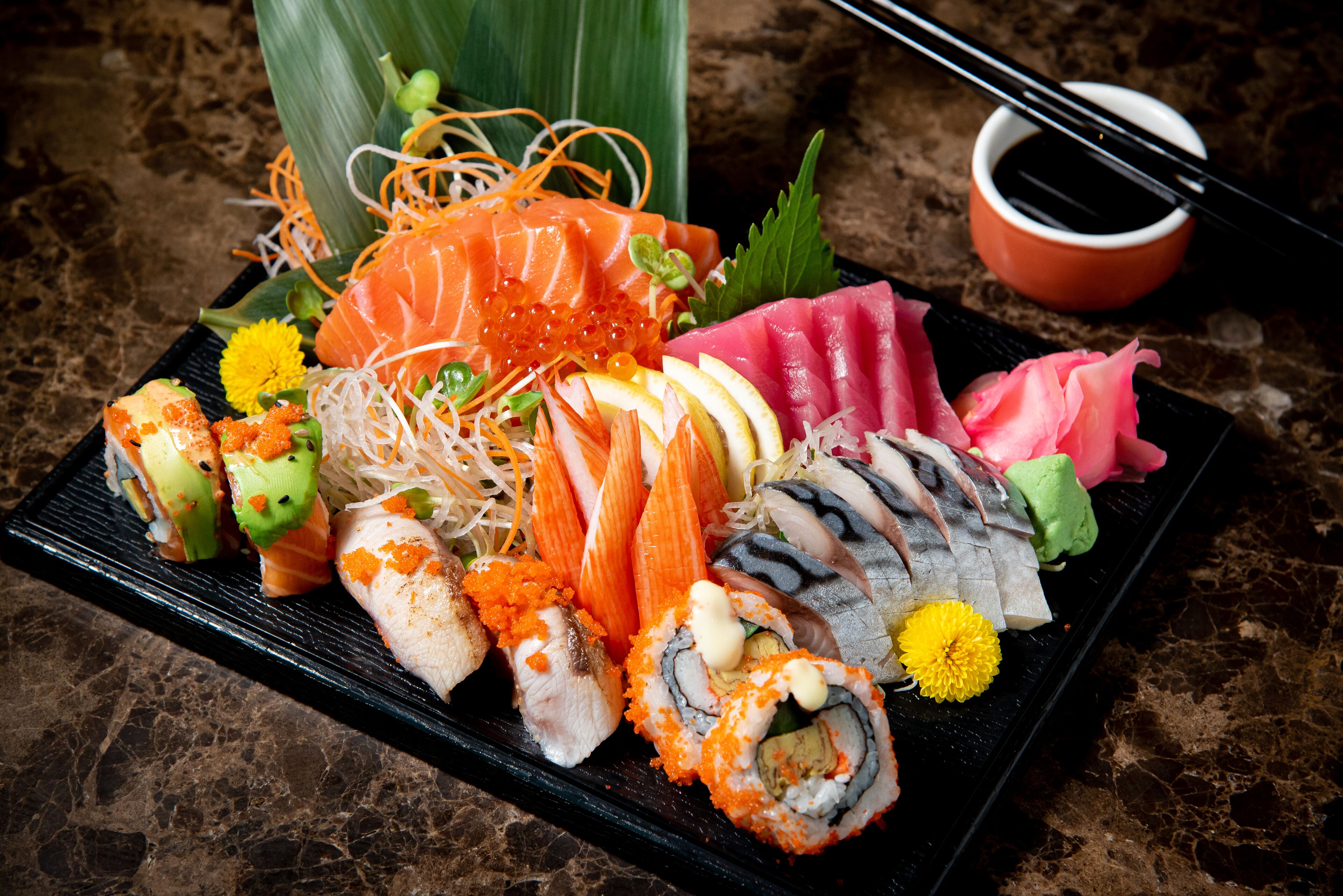 Verschiedene Sushi-Stücke auf einem schwarzen, rechteckigen Teller, dazu Stäbchen und eine kleine Sojasaucenschale.
