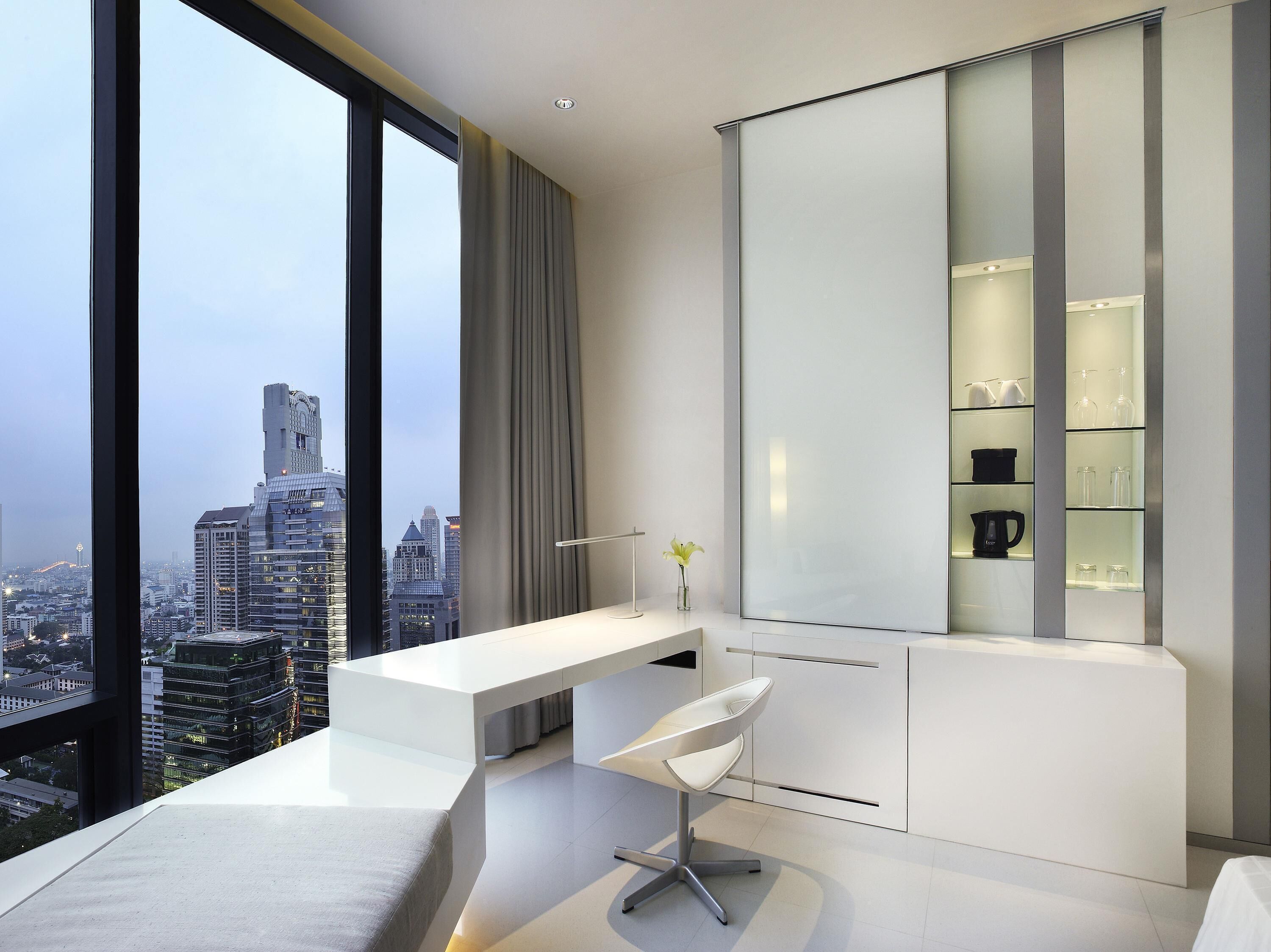 A modern hotel room with a large window, a white desk, a chair, and a built-in shelving unit.