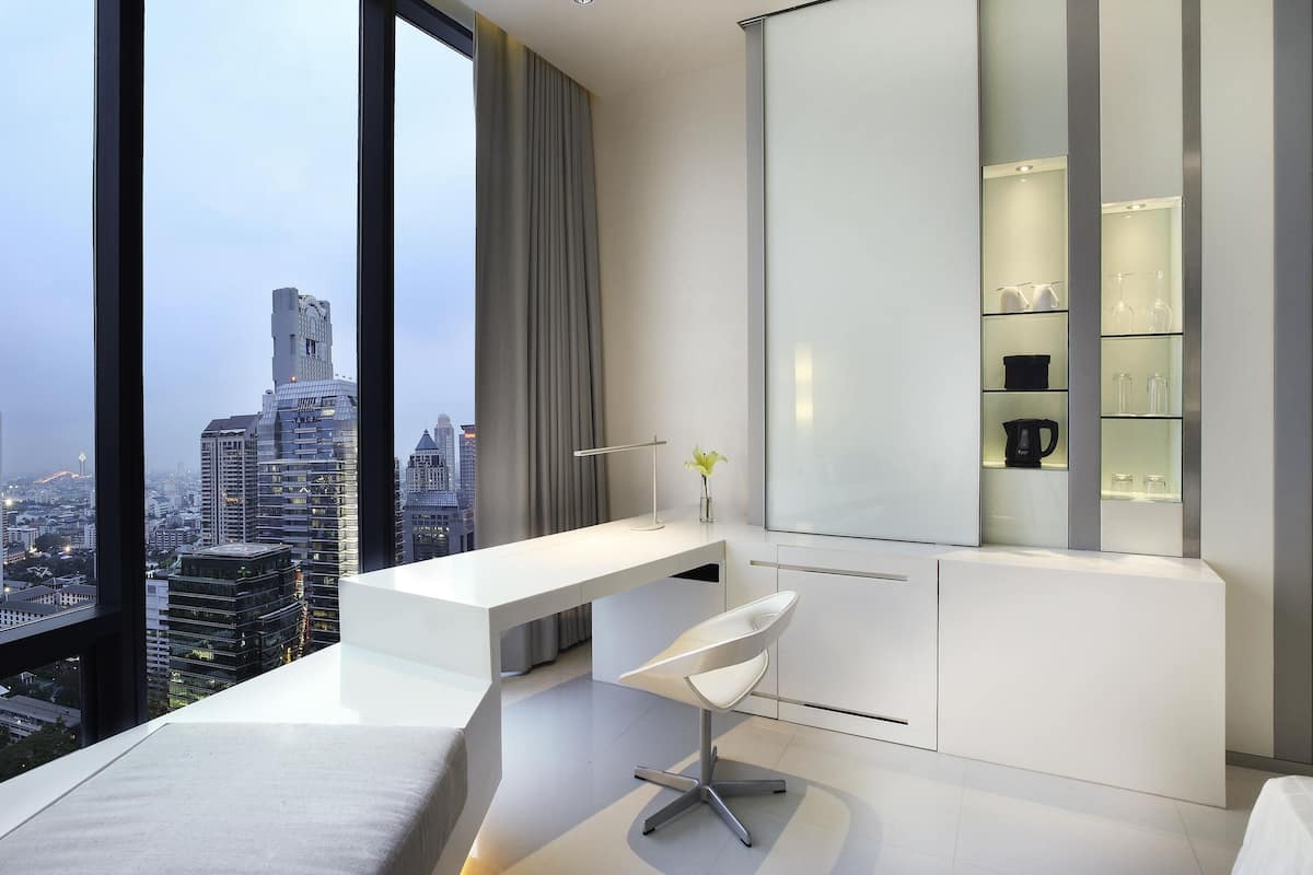A modern hotel room with a large window, a white desk, a chair, and a built-in shelving unit.
