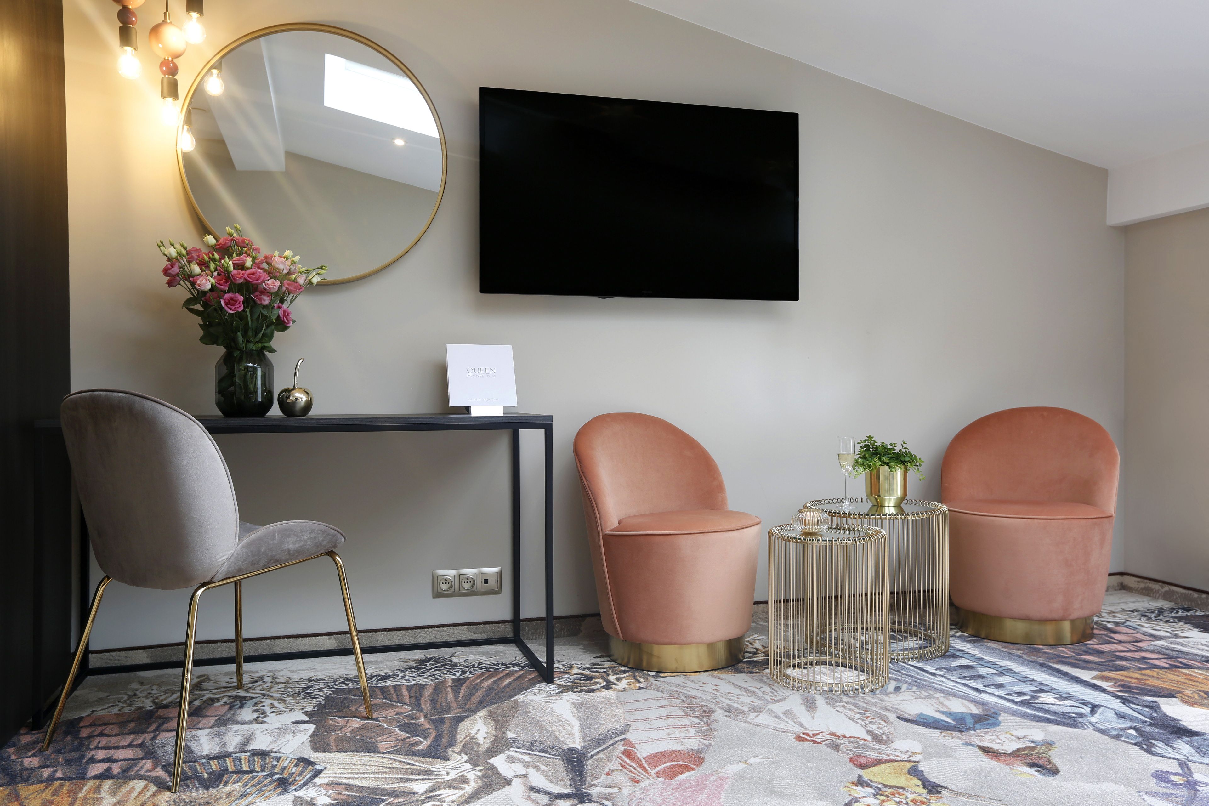 Ein modernes Hotelzimmer mit einem Flachbildfernseher, einem Schreibtisch mit Blumen in einer Vase, zwei rosafarbenen Sesseln, einem runden Beistelltisch mit einer Pflanze und einem gemusterten Teppich.