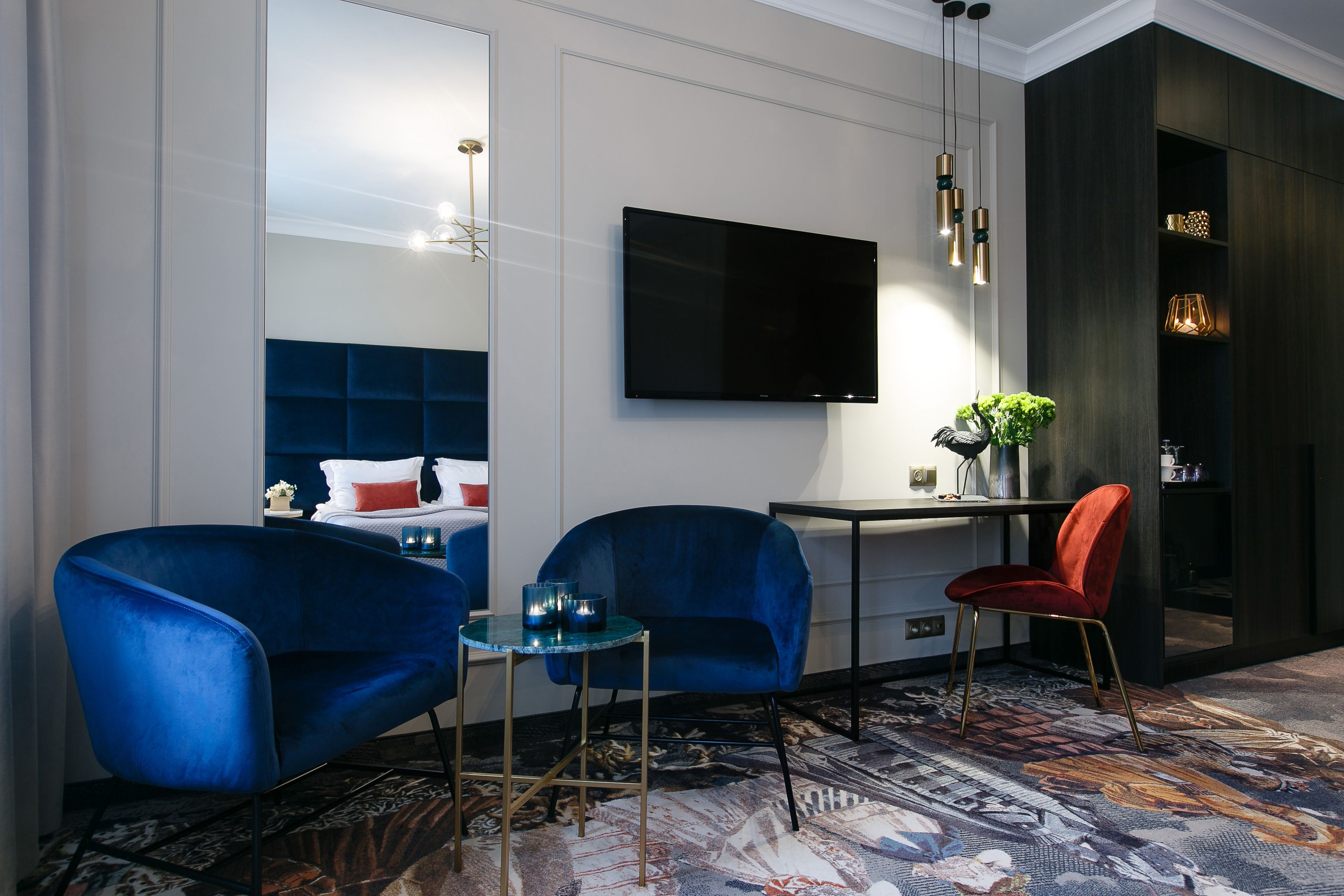 A modern hotel room with a large bed, two blue armchairs, a small table, a red chair, a TV mounted on the wall, and a patterned carpet.