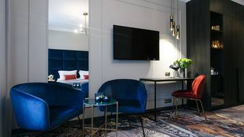 A modern hotel room with a large bed, two blue armchairs, a small table, a red chair, a TV mounted on the wall, and a patterned carpet.