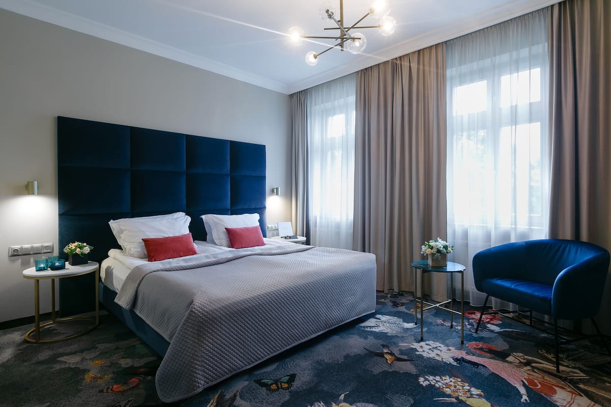 A modern hotel room with a large bed, a blue armchair, a small side table, and a decorative rug.