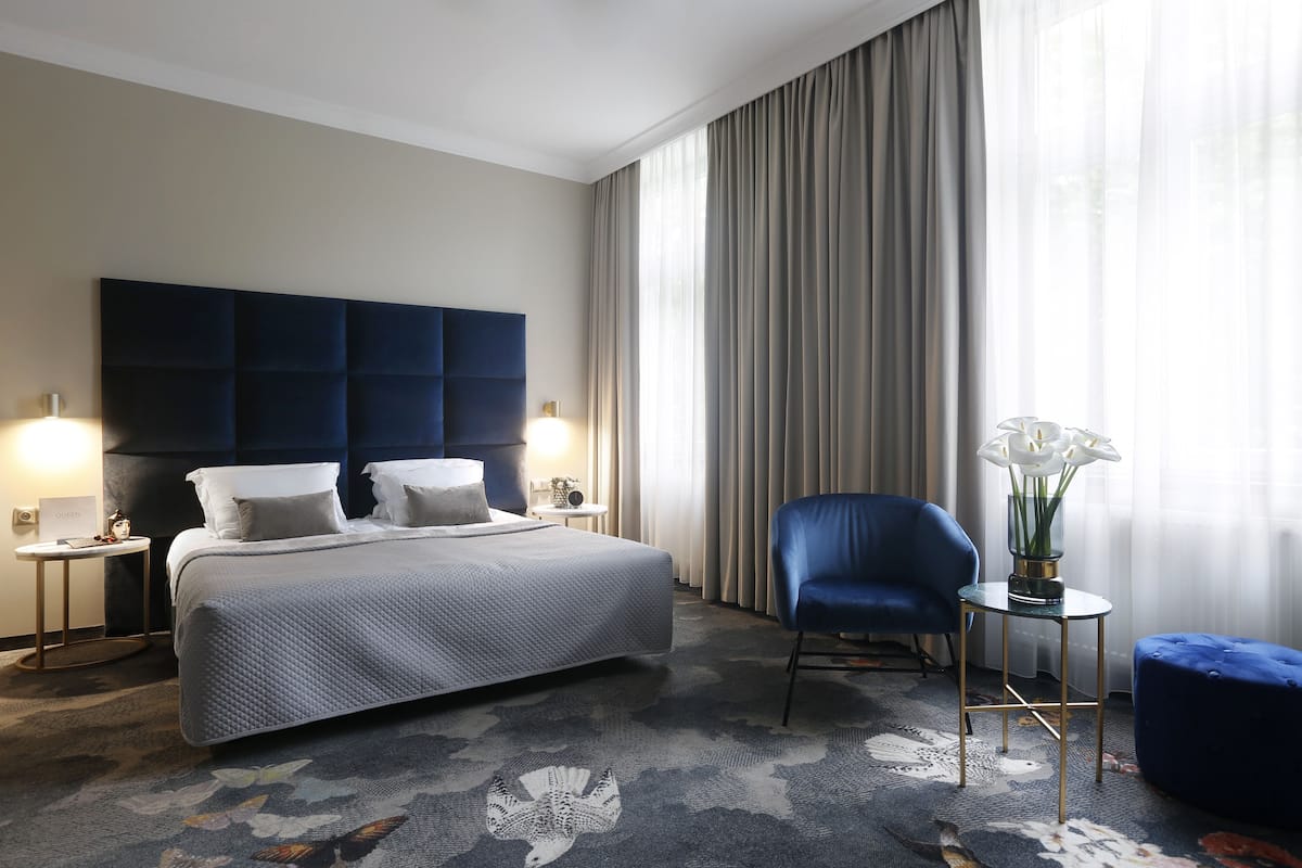 A hotel room with a large bed, two bedside tables with lamps, a blue armchair, a small side table with a vase, and a large window with curtains.