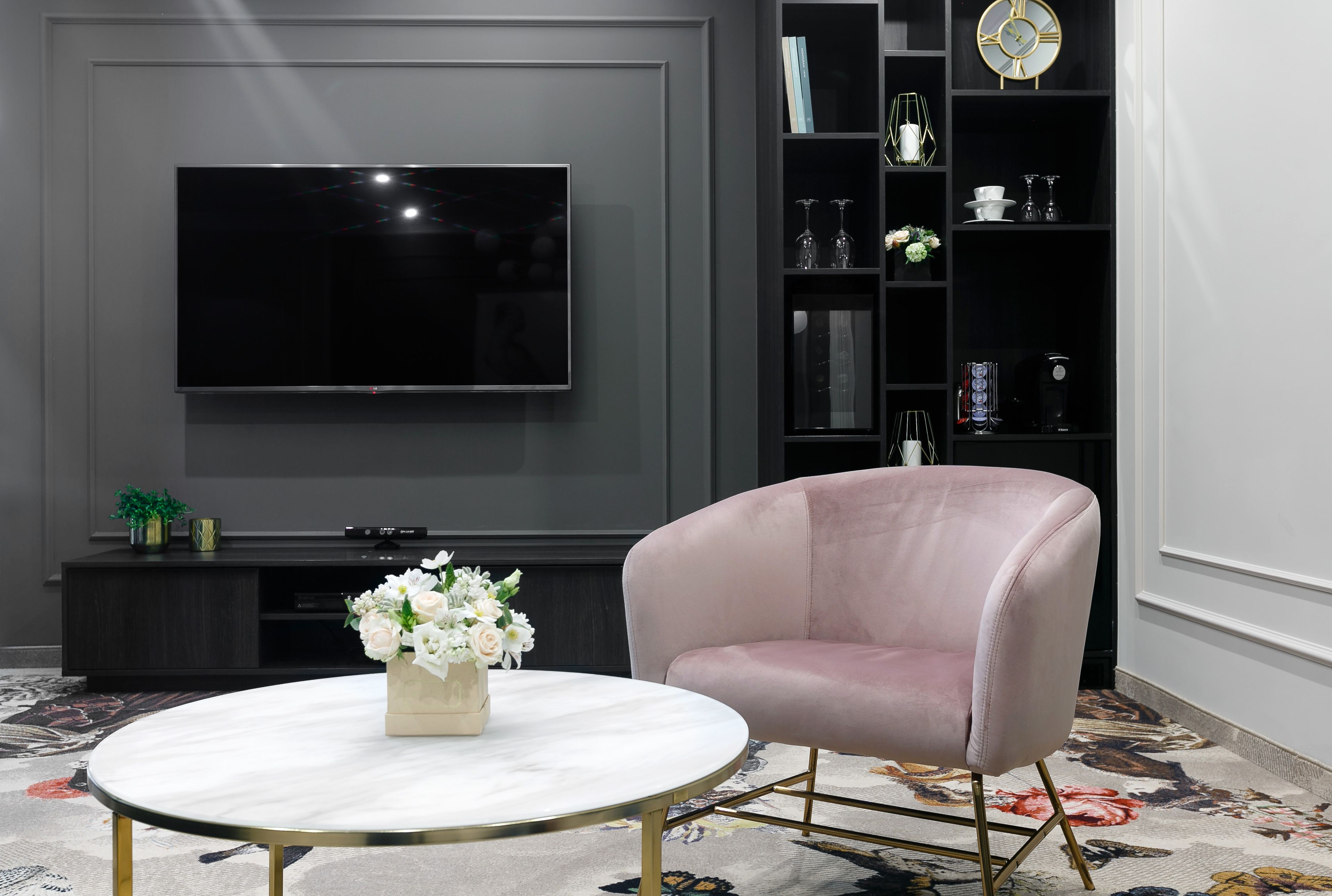 A modern living room with a flat-screen TV mounted on the wall, a round coffee table with a vase of flowers, a pink armchair, and a black entertainment unit with shelves.