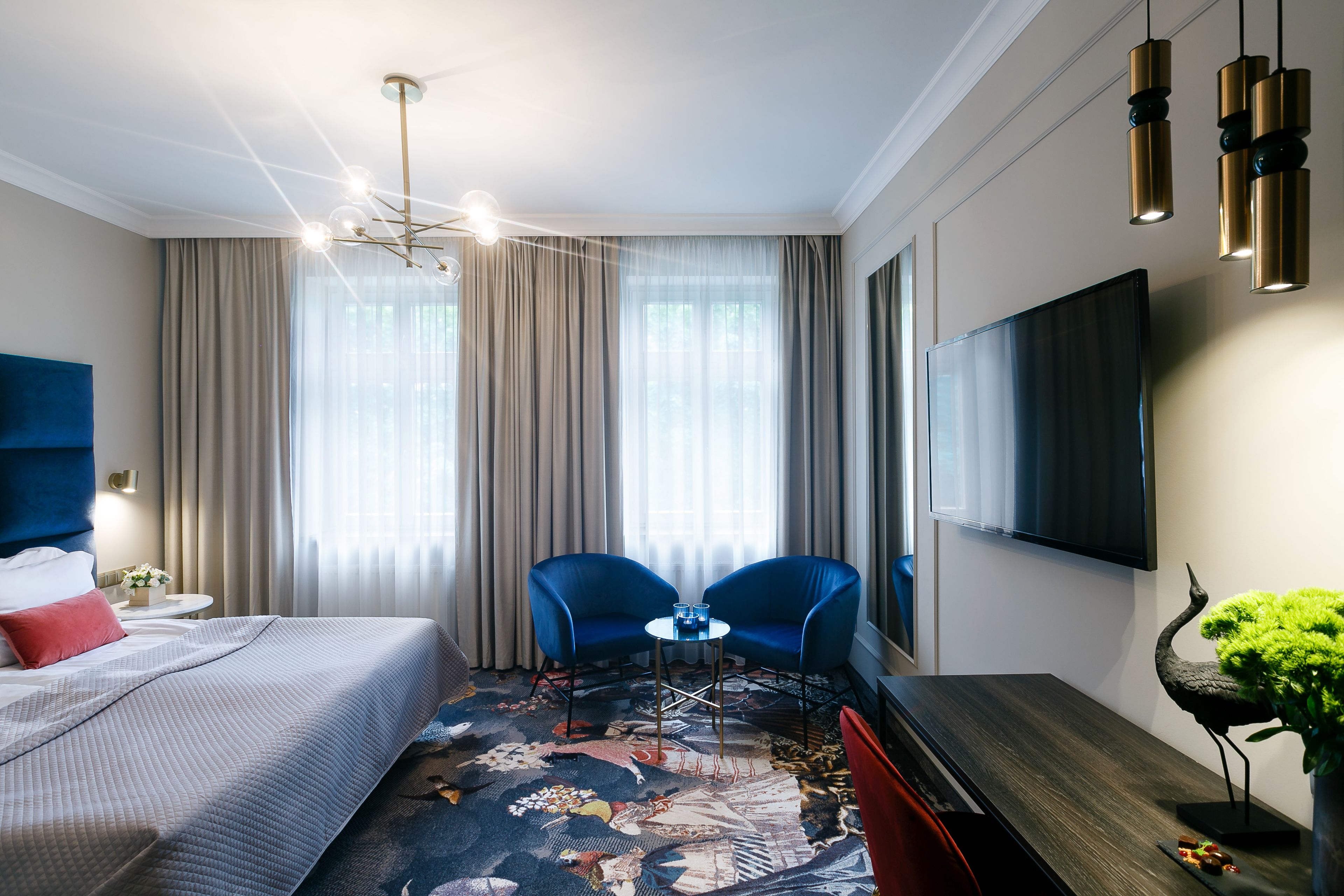 A hotel room with a bed, two blue chairs, a small table, a TV, and a decorative plant.