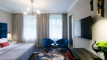 A hotel room with a bed, two blue chairs, a small table, a TV, and a decorative plant.