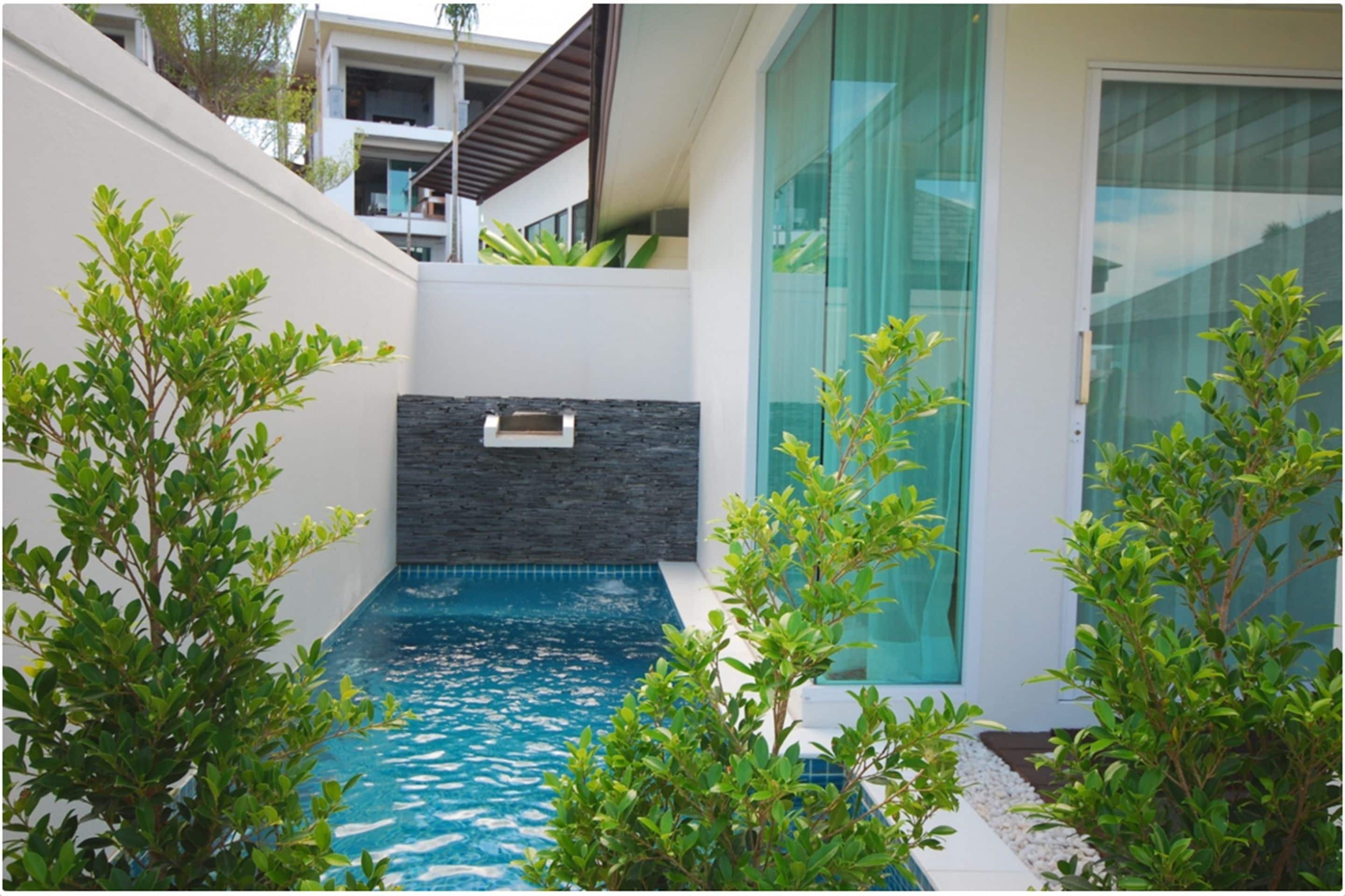 Un área de piscina exterior moderna con un muro de piedra, rodeada de vegetación y un ventanal.