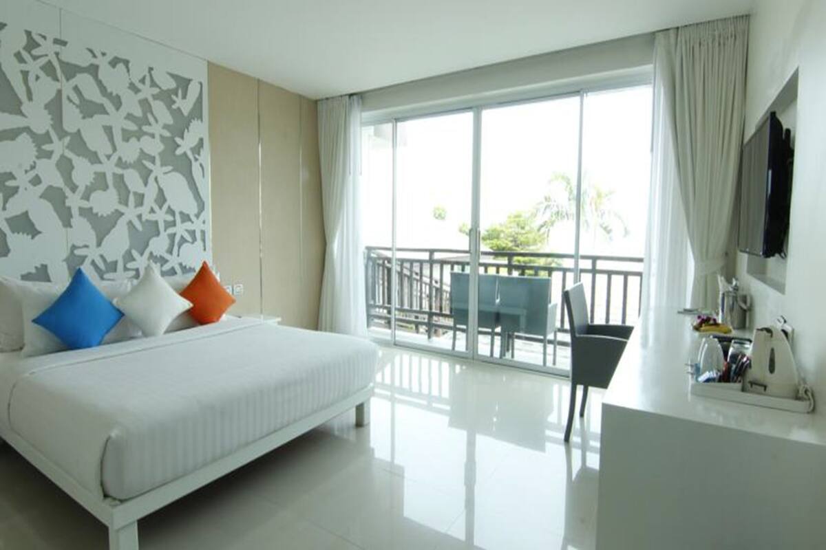 A modern hotel room with a large bed, a desk with a chair, and a balcony with a view of greenery.