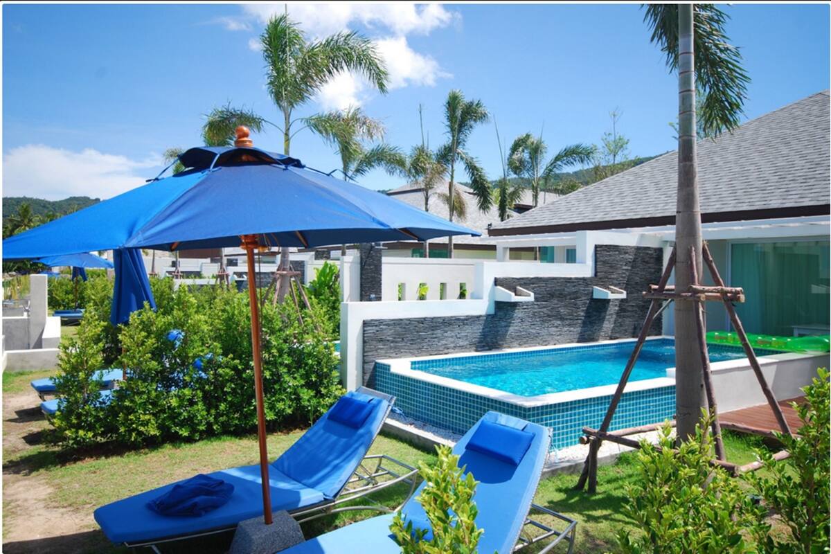 A swimming pool with lounge chairs and a blue umbrella, surrounded by greenery and palm trees.
