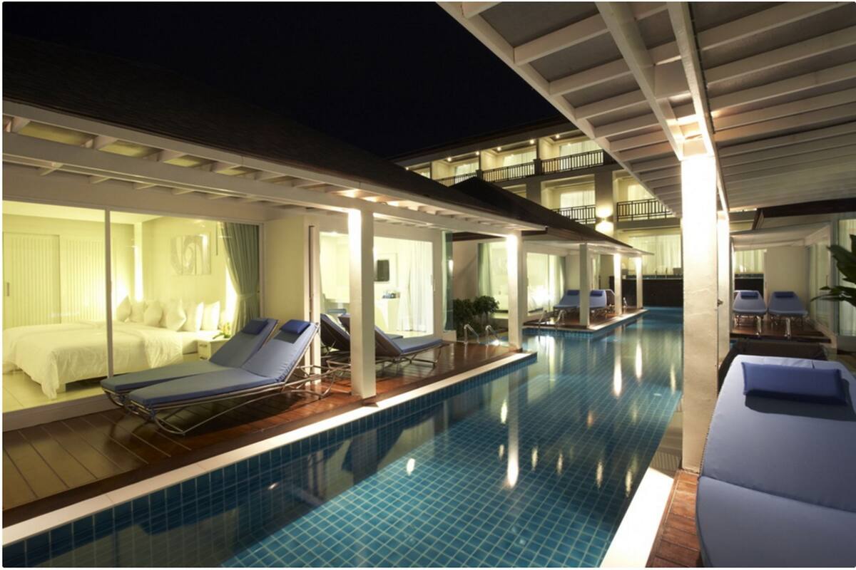 A swimming pool with lounge chairs and a bed, surrounded by a covered area with windows.