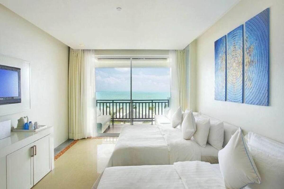A hotel room with two beds, a TV, and a balcony with a view of the ocean.