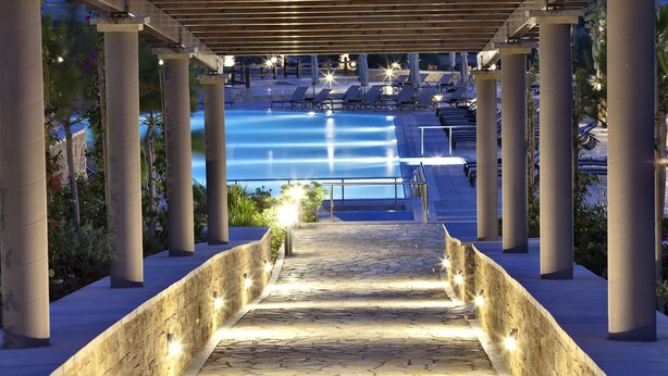 A stone pathway leading to a pool area under a pergola with columns, surrounded by lounge chairs and umbrellas.