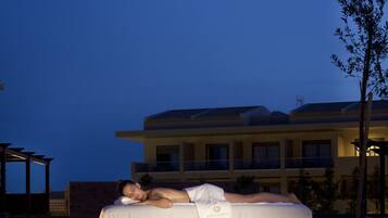 A person lying on a white-covered lounge chair by a pool at dusk.