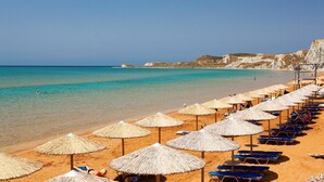 A beach with rows of sun umbrellas and lounge chairs, a clear blue sea, and rocky cliffs in the background.