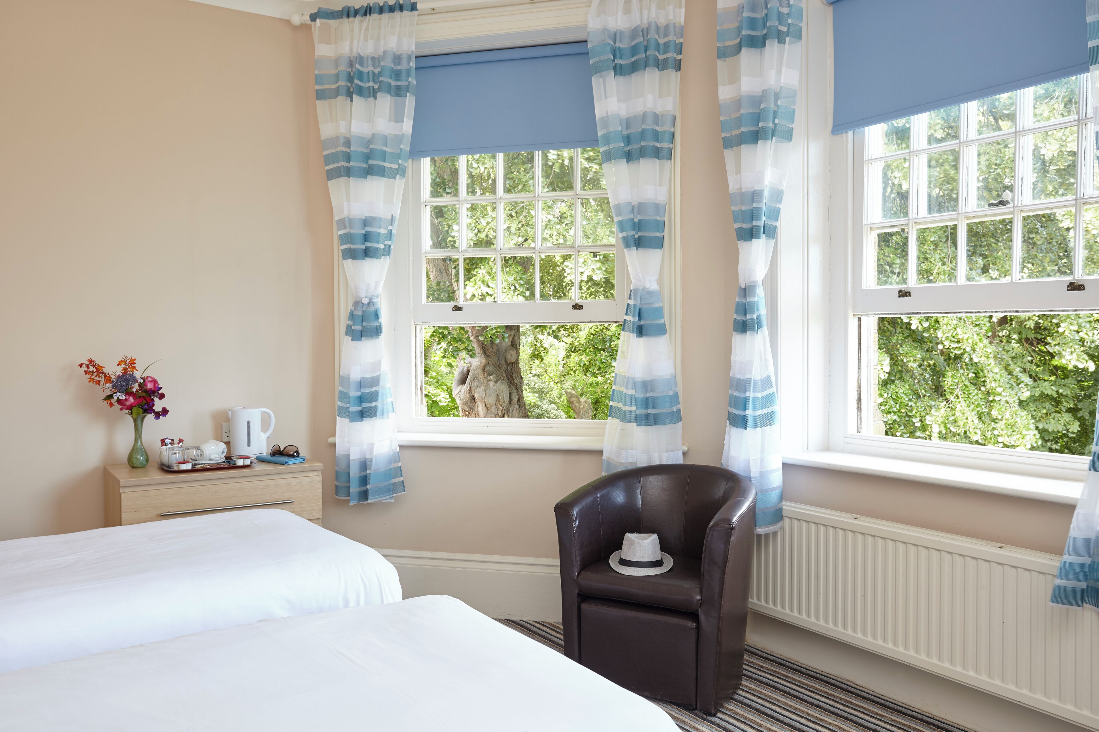 Ein Hotelzimmer mit zwei Betten, einem kleinen Tisch mit Blumenvase, einem Sessel und einem Fenster mit Vorhängen.