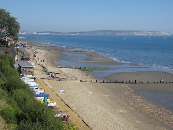 A coastal beach with a row of boats, a promenade, and a clear view of the sea and distant cliffs.