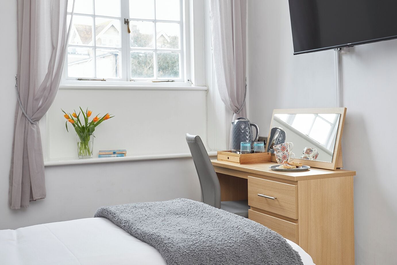 Un dormitorio con cama, escritorio con silla, ventana con cortinas, televisor de pantalla plana y un florero con flores.