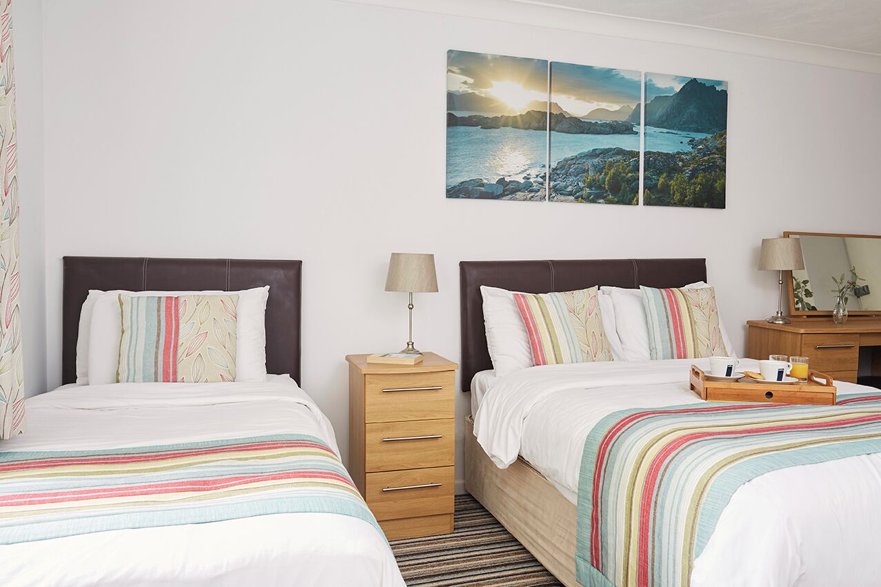 A hotel room with two beds, a wooden nightstand, a lamp, a tray with a cup and a glass, and a painting of a coastal landscape.