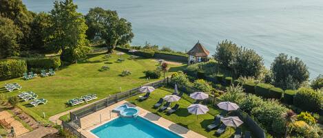 A pool area with lounge chairs and umbrellas, surrounded by well-maintained lawns and a view of the sea.