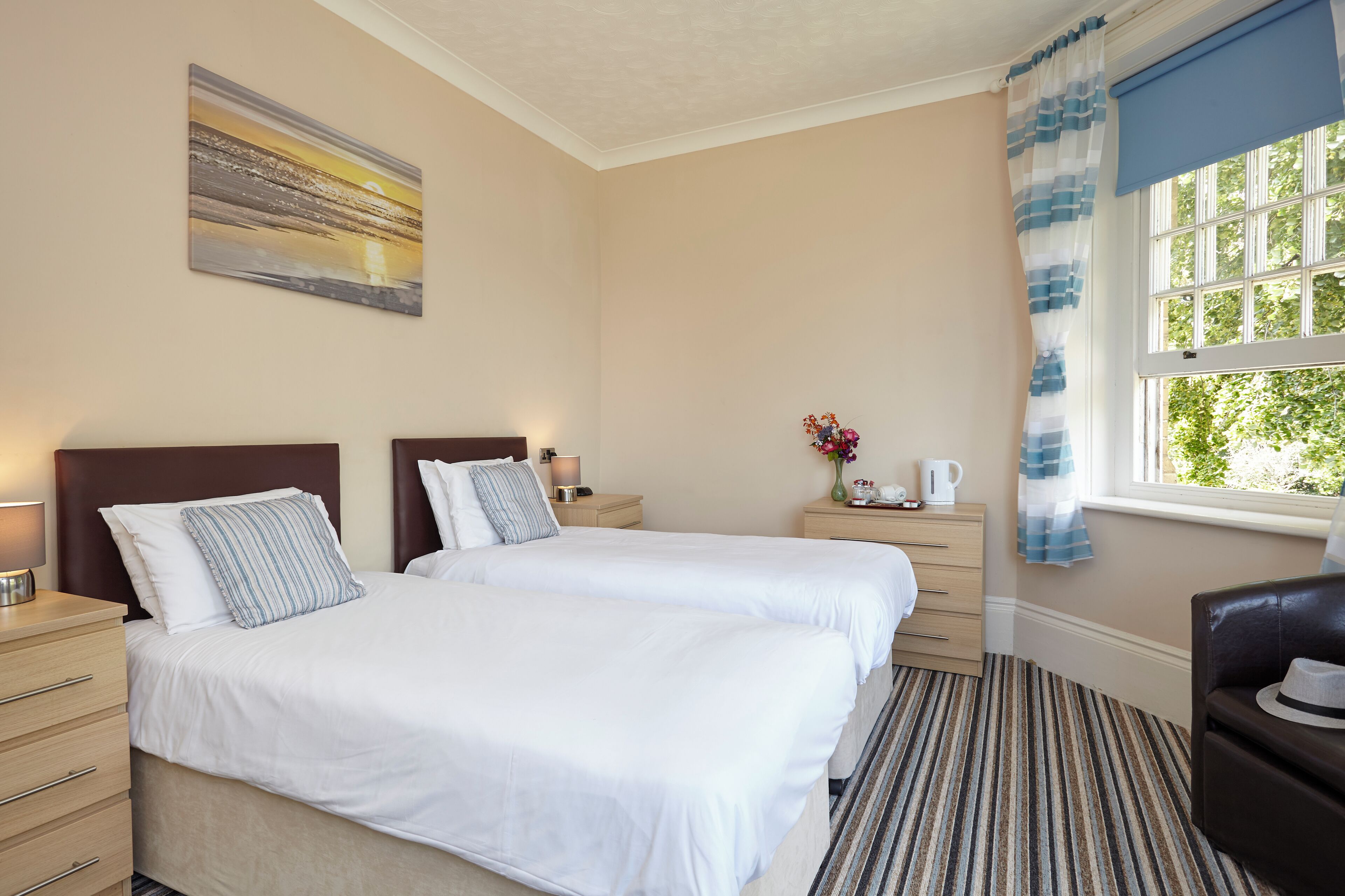 Une chambre d’hôtel avec deux lits, une table de chevet, une lampe, un vase avec des fleurs, une machine à café et une vue sur la verdure par la fenêtre.