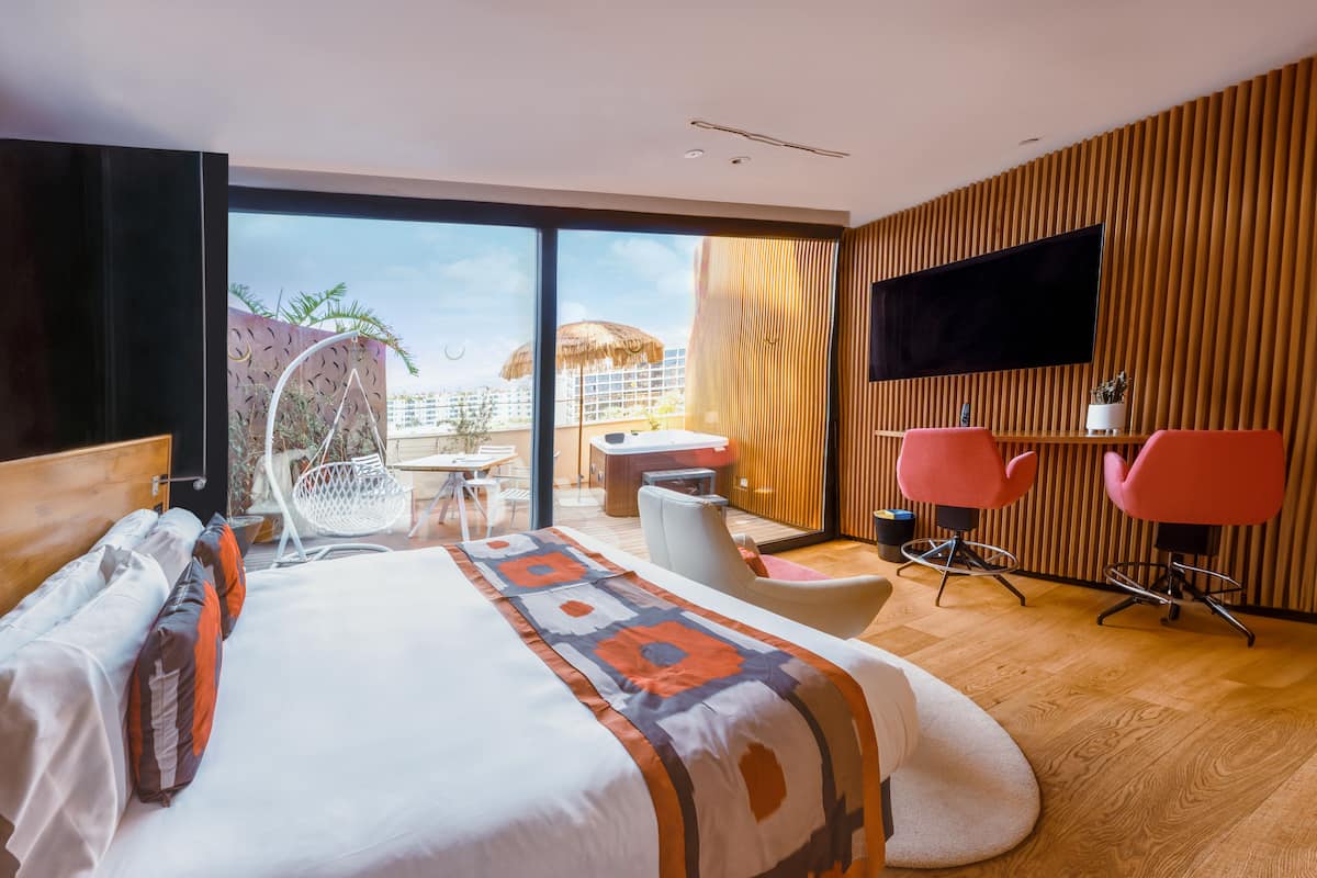 A modern hotel room with a large bed, a desk, two pink chairs, a TV, and a balcony with a view of buildings and palm trees.