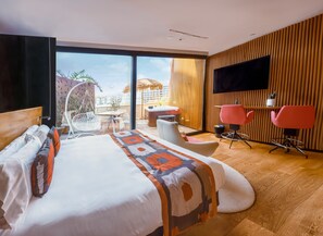 A modern hotel room with a large bed, a desk, two pink chairs, a TV, and a balcony with a view of buildings and palm trees. - Bohemia Suites & Spa - Adults only (San Bartolomé de Tirajana)