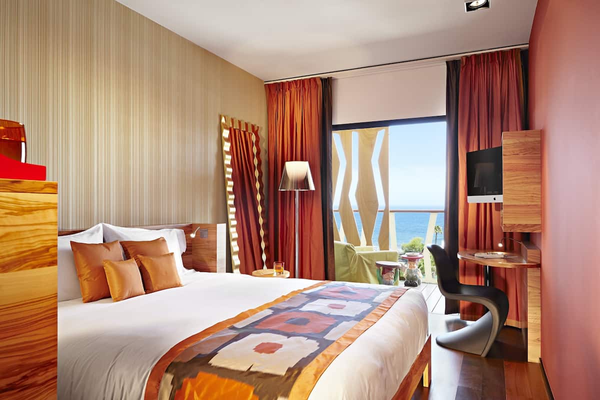A hotel room with a large bed, a desk, a chair, a TV, and a view of the ocean.