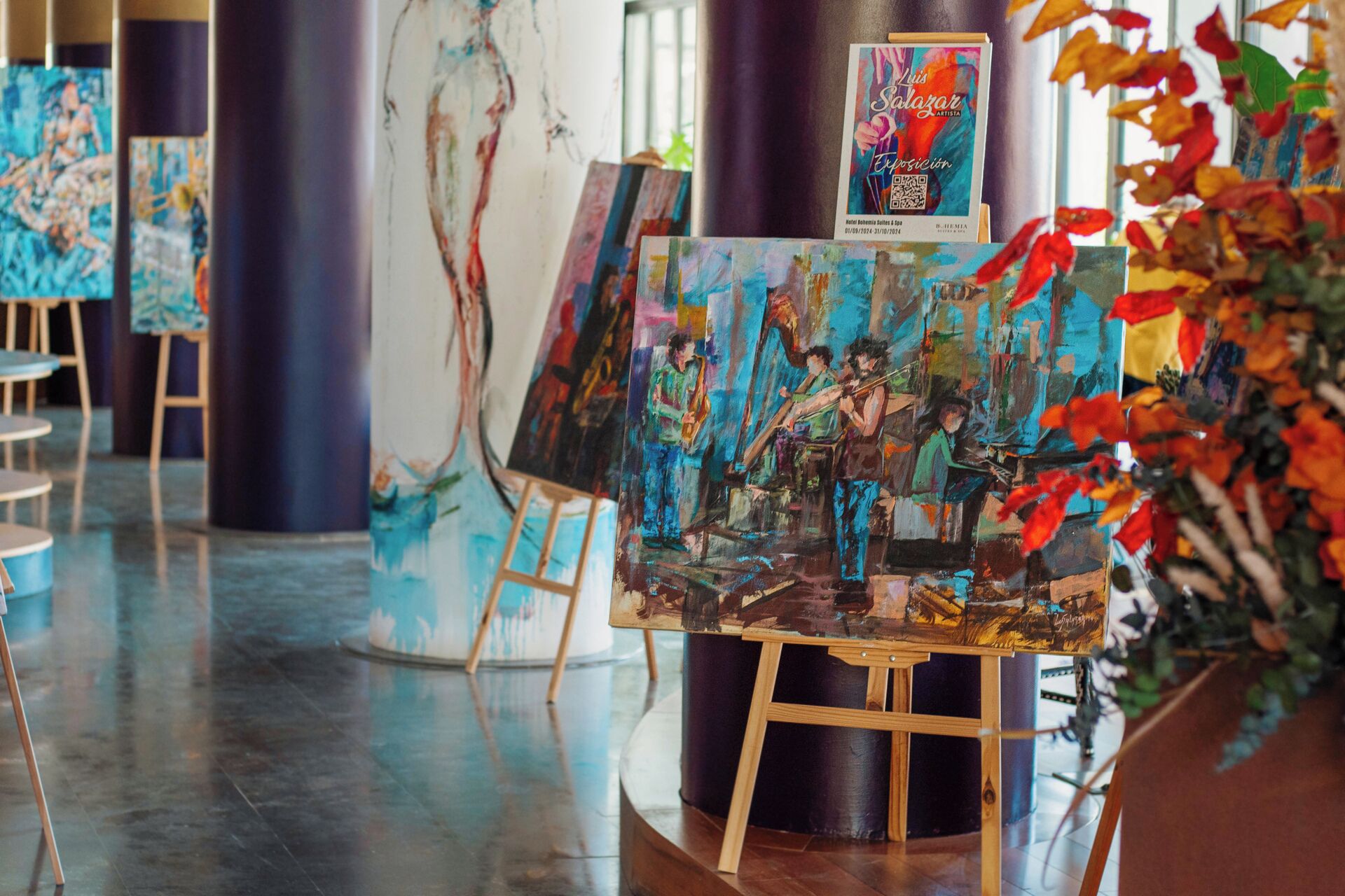 Una galería de arte con pinturas sobre caballetes, un cartel y una planta en maceta con hojas otoñales.
