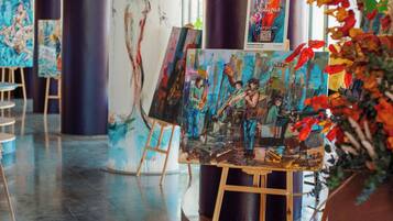 Una galería de arte con pinturas sobre caballetes, un cartel y una planta en maceta con hojas otoñales.