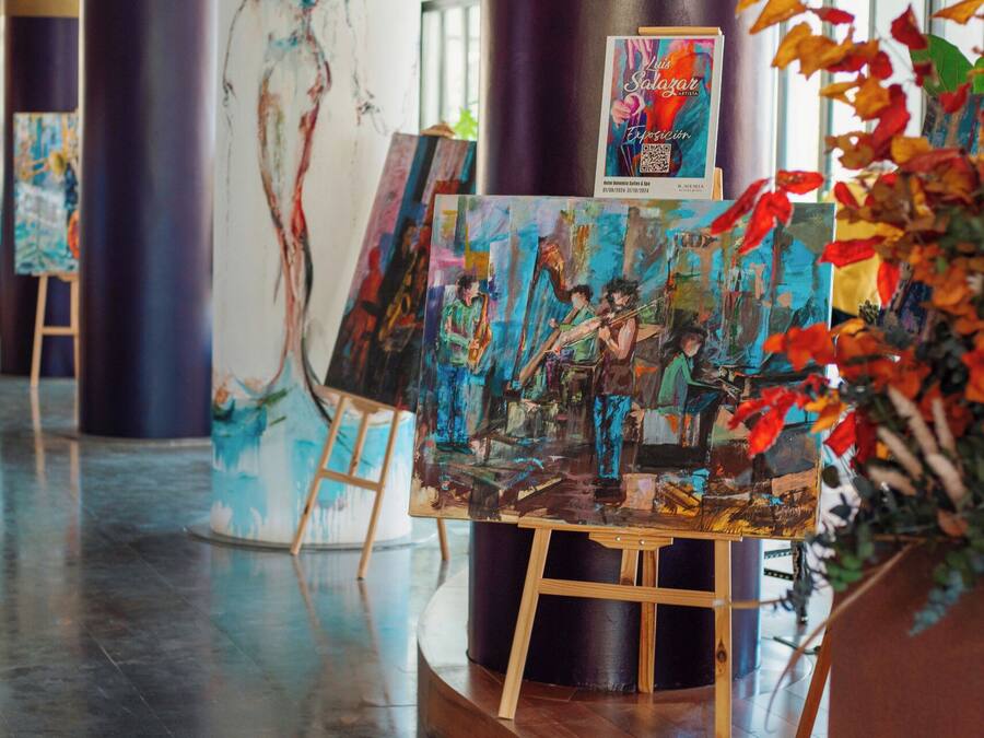 Una galería de arte con pinturas sobre caballetes, un cartel y una planta en maceta con hojas otoñales.