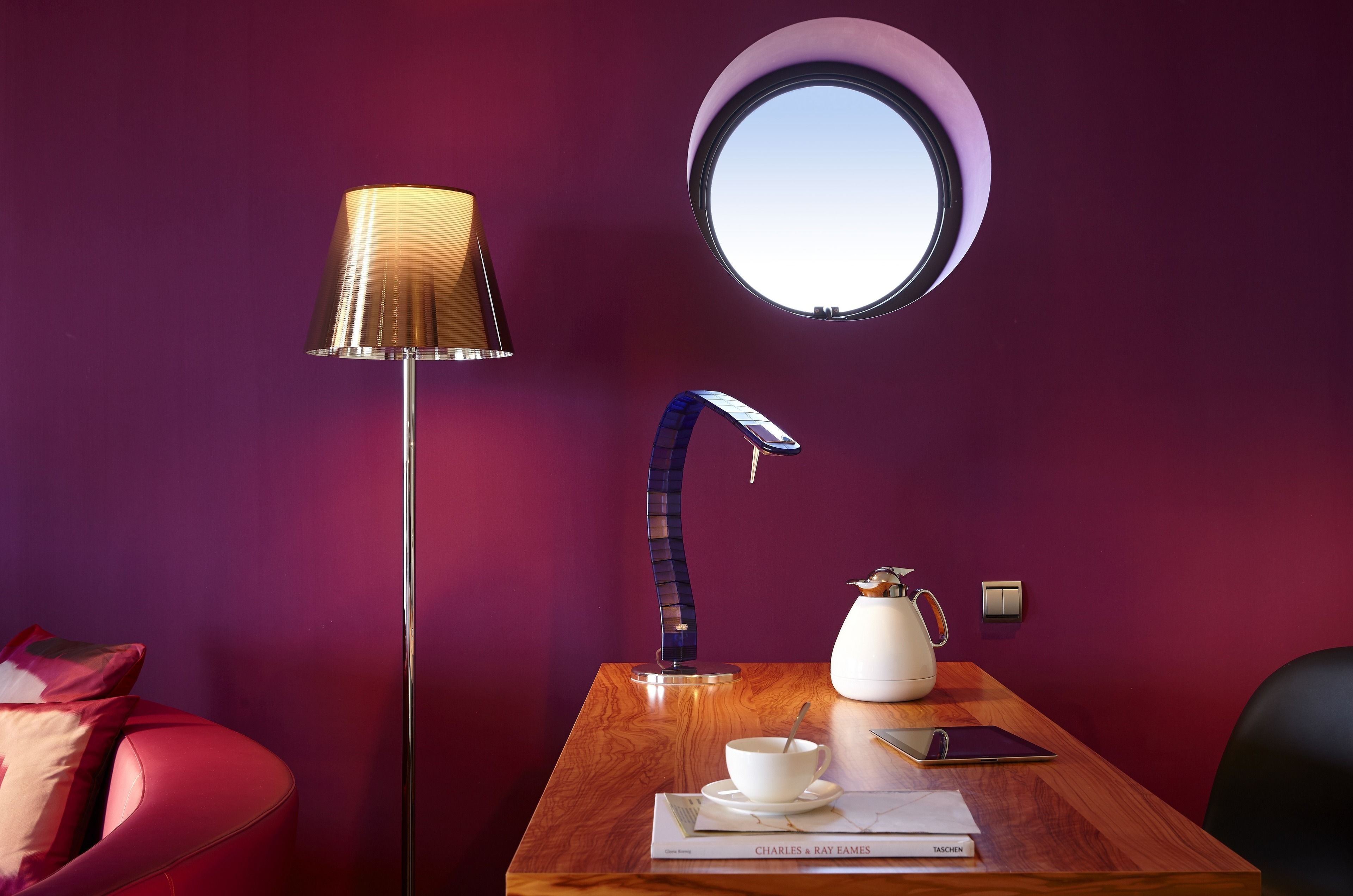 Una habitación con un ventanal redondo, una lámpara moderna, una mesa con un hervidor de agua, una taza y un libro titulado "Charles & Ray Eames".