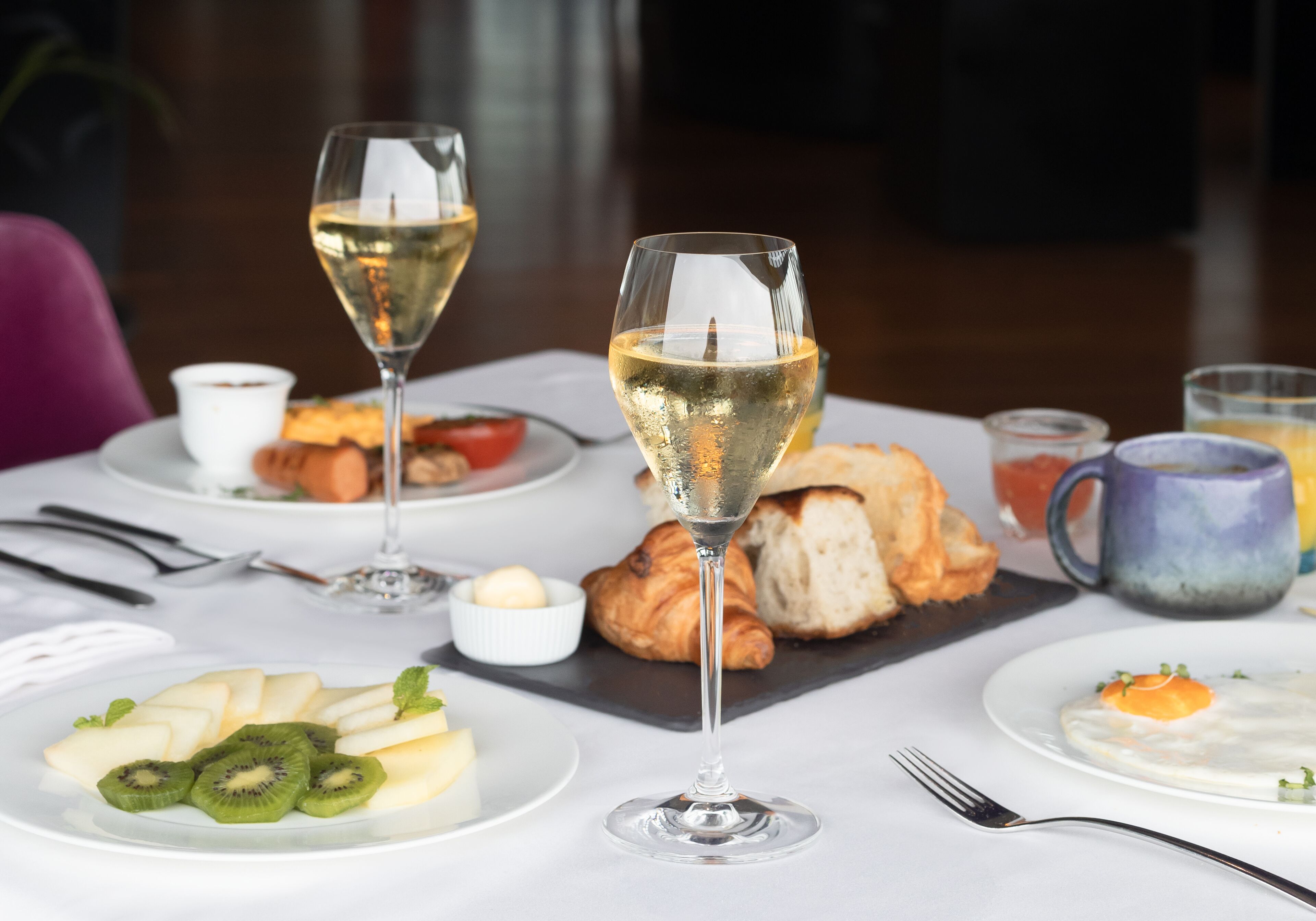 A dining table with a croissant, a glass of white wine, and plates of food including kiwi slices and a poached egg.