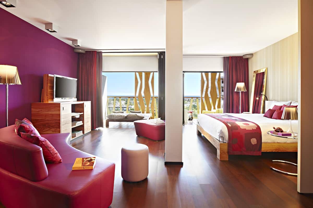 A modern hotel room with a large bed, a sofa, a TV, and a balcony with a view.