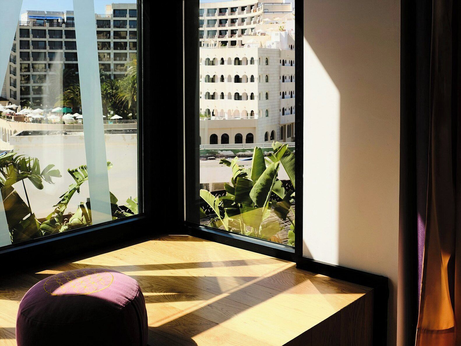 Una habitación con un ventanal grande que da a un paisaje urbano, un banco de madera y vistas a edificios y palmeras.