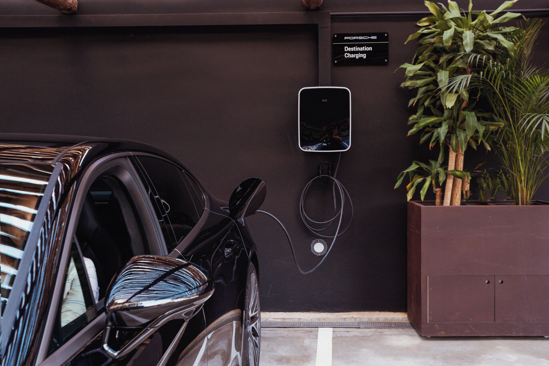 A car parked in a garage with a charging station, a sign indicating 'Destination Charging,' and a potted plant beside the wall.