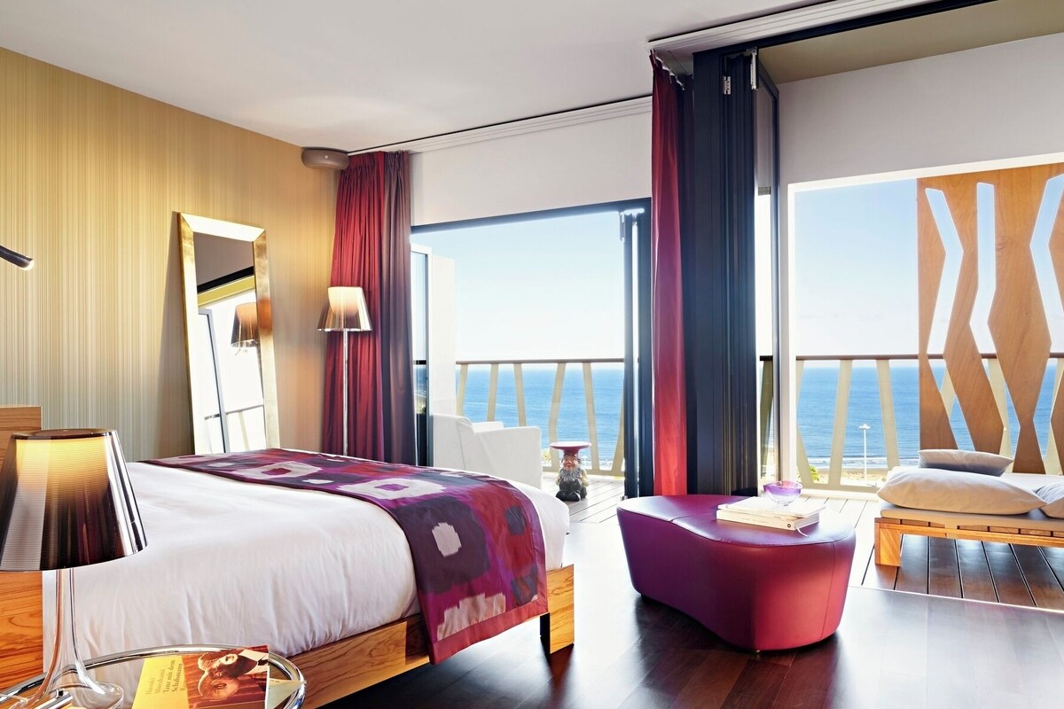 Una habitación de hotel moderna con una cama grande, un reposapiés rojo, un balcón con vistas al mar y un ventanal amplio.