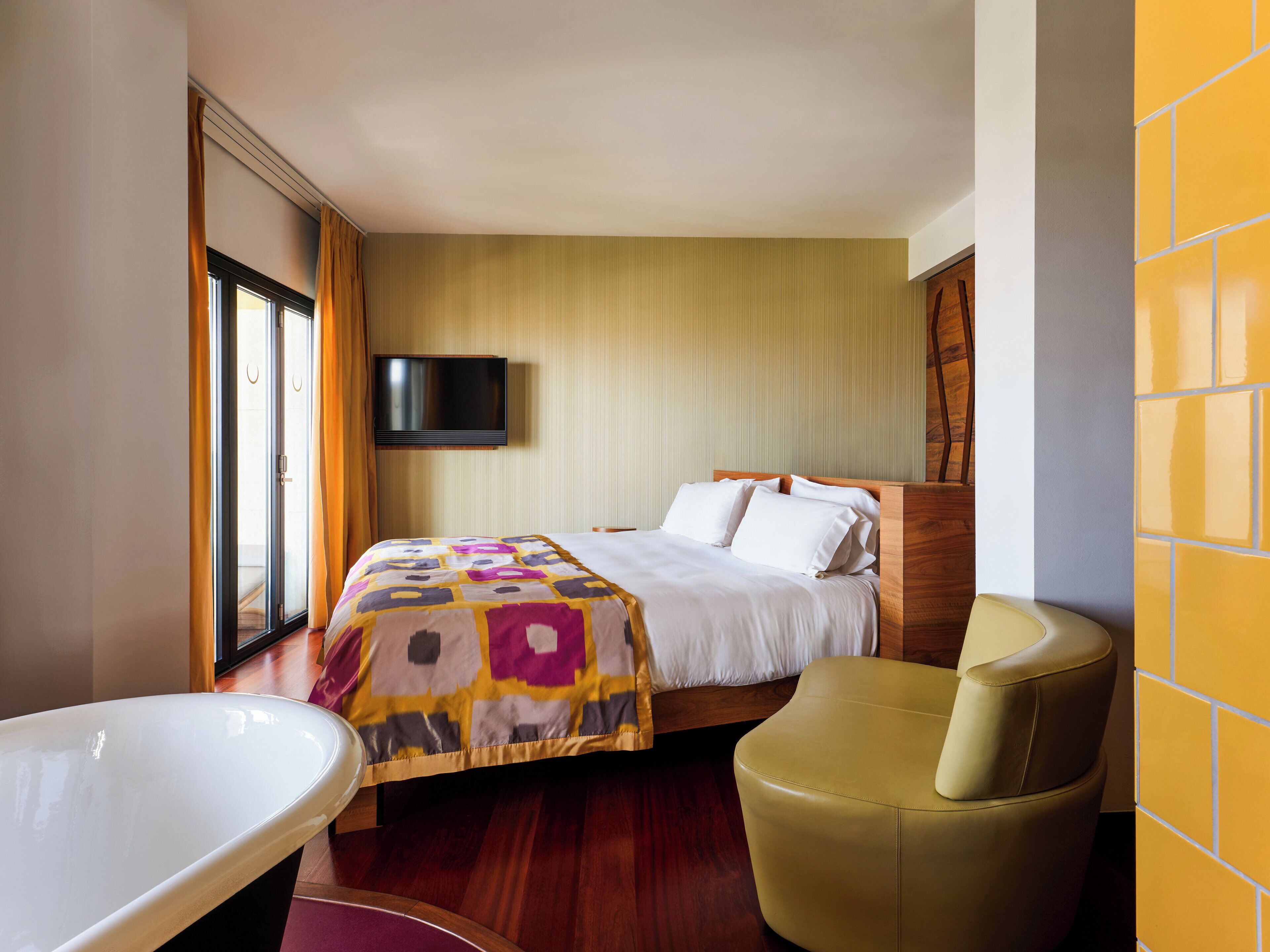 A hotel room with a bed, a television mounted on the wall, a bathtub, and a chair.