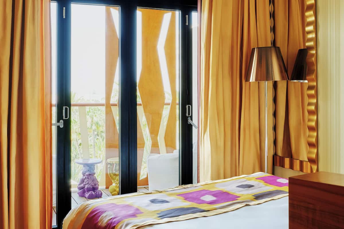 A hotel room with a bed, a round wooden table, a lamp, and curtains.